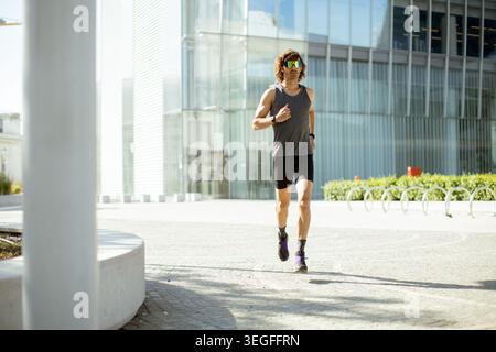 Eine Person läuft auf einem gepflasterten Weg in einem städtischen Gebiet. Der Läufer trägt eine Sonnenbrille und Sportausrüstung. Helles Sonnenlicht scheint auf die Szene und hebt einen Mod hervor Stockfoto