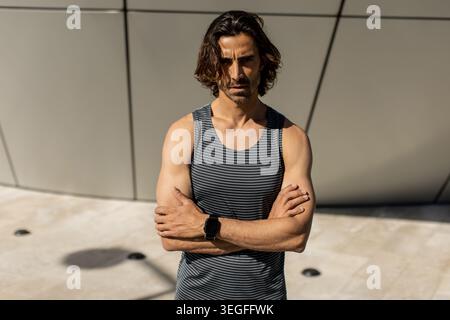 Ein Mann mit langen Haaren steht mit überkreuzten Armen. Er trägt ein ärmelloses Hemd. Die Umgebung ist hell und es gibt ein modernes Gebäude im Hinterkopf Stockfoto