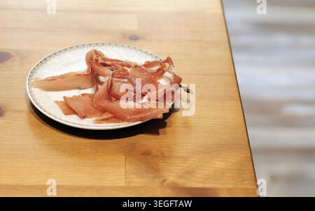 Dünn geschnittener geräucherter Schinken, mit schwarzem Pfeffer abgeschmeckt, auf einem dekorativen Teller präsentiert, auf einem warmen Holztisch für eine köstliche Vorspeise oder sna Stockfoto