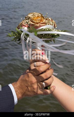 Bräutigam und Braut halten einen Hochzeitsstrauß mit goldenen Rosen und Ringen, die die Ehe symbolisieren Stockfoto