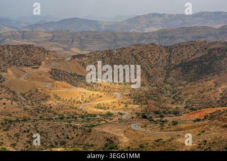 Eine asphaltierte Straße schlängelt sich durch eine trockene Berglandschaft in der Region Afar, Äthiopien. Das Sonnenlicht beleuchtet felsige Hügel und spärliches Gestrüpp und hebt die r Stockfoto