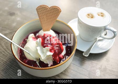 Eisbecher, Schaufeln Vanilleeis mit Schlagsahne und rotem Fruchtkompott in einer Schüssel und eine Tasse Cappuccino auf einem Holztisch, Sommerdessert, Sel Stockfoto