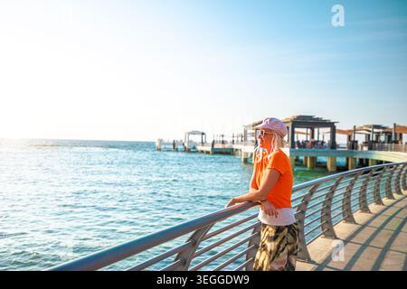 Blonde Frau mit Hut und orangenem T-Shirt, die sich an ein Geländer lehnt und den Blick auf das Rote Meer und die Uferpromenade von Jeddah mit Wellen genießt Stockfoto