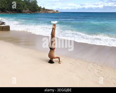 Anmutige schlanke Frau, die einen Kopfstand an einem unberührten tropischen Strand mit weichem weißem Sand und kristallklarem türkisfarbenem Wasser übt. Von hinten gesehen. Stockfoto