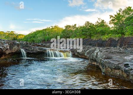 Eine kaskadierende Wasserquelle fließt über felsiges Gelände, umgeben von lebendigen grünen Pflanzen unter einem klaren blauen Himmel in Guaviare. Stockfoto