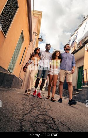 Niedriger Blickwinkel auf verschiedene junge Freunde, die gemeinsam durch eine farbenfrohe mediterrane Straße spazieren, Freundschaft genießen und Sommerreisen genießen, während sie erkunden Stockfoto