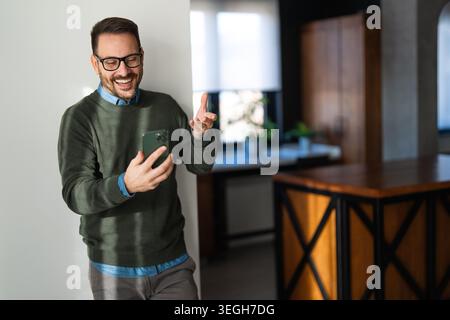 Selbstbewusster, lächelnder Geschäftsmann mit Smartphone im Heimbüro. Überprüfen neuer E-Mails, Videoanrufe Stockfoto