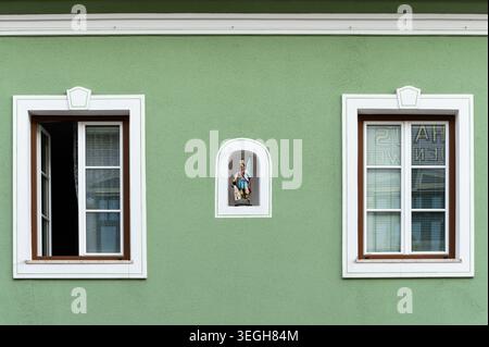 Mariazell, Österreich - 12. Juni 2020: Kleine gemalte heiligenfigur-Schrein-Nische zwischen weiß gerahmten Fenstern auf verwitterter grüner Stuckfassade mit Reflexion Stockfoto