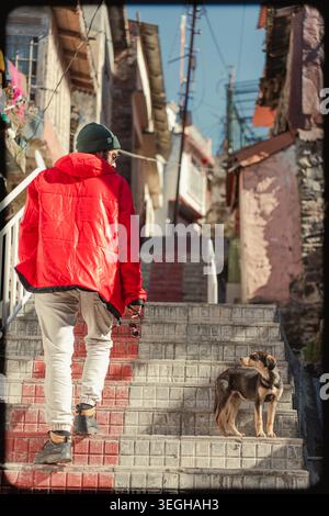 Eine Person, die eine leuchtend rote Jacke trägt, geht in Begleitung eines kleinen Hundes in Chakrata, Uttarakhand, Indien, eine Steintreppe hinauf. Stockfoto