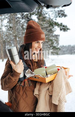 Die Frau hält Getränke und warme Kleidung, während sie im Schnee steht Stockfoto