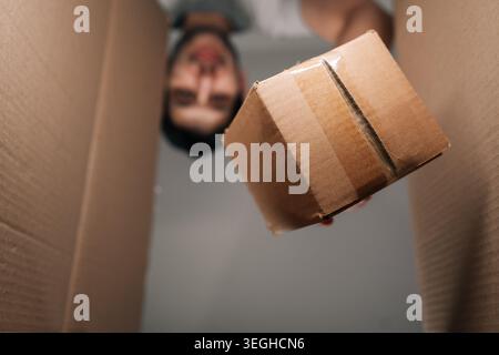 POV-Aufnahme mit niedrigem Winkel aus der Pappschachtel. Ein Mann mit verschwommenem Gesicht legt ein kleineres Paket hinein. Konzept für Transport, Versand, Lieferung, E-Commerce Stockfoto