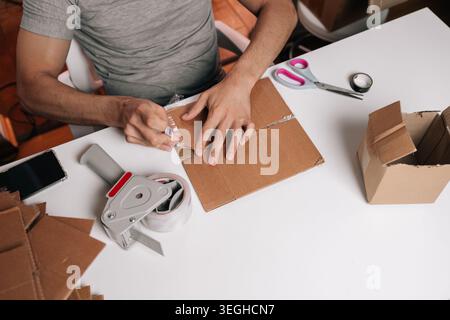 Hochwinkelansicht der Person, die Pappkarton verpackt. Der Eigentümer eines kleinen Unternehmens bereitet das Paket für den Versand vor. Konzept für E-Commerce, Fulfillment, Delivery Stockfoto