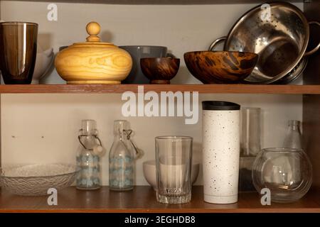 Eine Sammlung verschiedener Küchenutensilien, darunter Holzschalen, Glasbecher, Flaschen und ein Sieb, wird ordentlich in rustikalen offenen Regalen gelagert. Stockfoto
