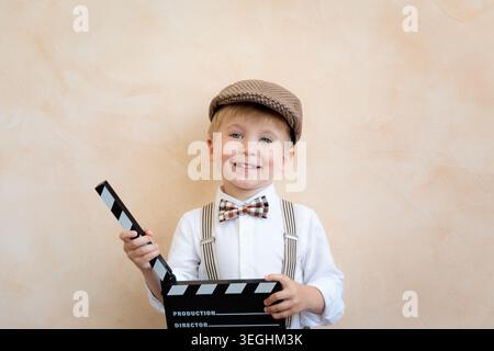 Lustiger Junge, der ein Klappbrett hält. Glückliches Kind, das zu Hause Spaß hat. Retro-Kino-Konzept Stockfoto