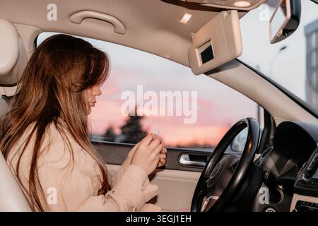 Frau Auto Make-up junge Frau, die auf dem Fahrersitz eines Fahrzeugs sitzt und Lippenstift während eines schönen rosa Sonnenuntergangs aufträgt Stockfoto