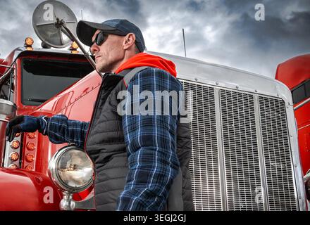 Ein Mann mit Mütze und Sonnenbrille steht neben einem roten Truck. Er trägt ein kariertes Hemd und eine Weste. Der Stapler verfügt über einen großen Frontgrill und Scheinwerfer. Der Himmel Stockfoto