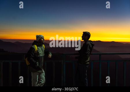 Silhouetten von zwei Wanderern, die auf einem Kamm stehen und den lebhaften Sonnenuntergang über den nebeligen Himalaya-Tälern in Chakrata, Uttarakhand, Indien, bewundern. Stockfoto