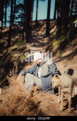 Eine junge Frau mit Winterjacke und Mütze sitzt mit ihrem Hund auf einem Waldweg in den malerischen Bergen von Chakrata, Uttarakhand, Indien. Stockfoto