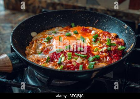 Eine Nahaufnahme eines lebhaften Eiergerichts, das in einer Pfanne zubereitet wird, garniert mit frischen grünen Zwiebeln und Tomaten, wodurch eine farbenfrohe und appetitliche Mahlzeit entsteht. Stockfoto
