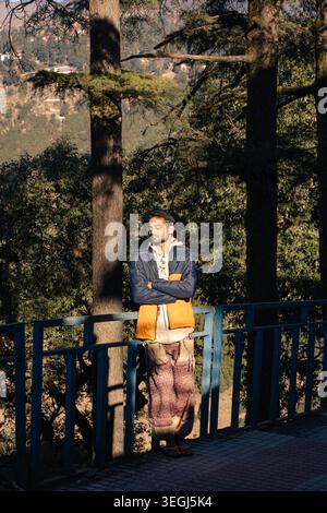 Ein junger Mann steht mit gekreuzten Armen an einem blauen Geländer in einem sonnendurchfluteten Kiefernwald an einem friedlichen Nachmittag in Chakrata, Uttarakhand, Indien. Stockfoto
