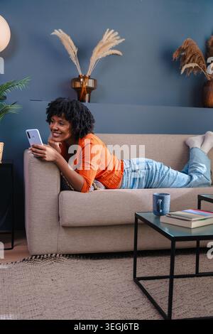 Vertikale Portrait afroamerikanerin, die auf dem Sofa liegt und zu Hause ihr Smartphone benutzt Stockfoto