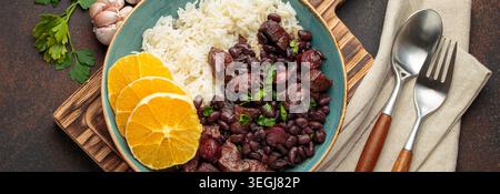 Herzhafter brasilianischer Feijoada-Eintopf mit schwarzen Bohnen, Schweinefleisch, Rindfleisch, Reis und Orangenscheiben in einer rustikalen Schüssel auf Holzbrett mit Besteck und Serviette. Traditionelles Gericht aus Brasilien Stockfoto