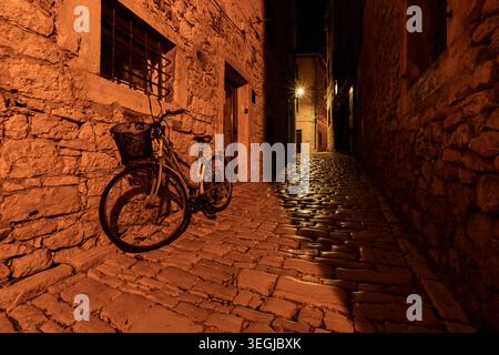 Abgestelltes Fahrrad mit Korb auf einer kopfsteingepflasterten Straße in einer engen Gasse der Altstadt von Rovinj, Kroatien bei Nacht. Stockfoto