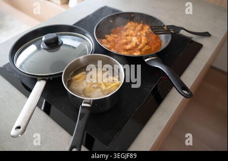Zwei Pfannen, die Pasta und Soße auf einem Induktionskochofen kochen, Tortellini in einem Topf, knallendes Tomaten-Hühnchen-Ragu in einer Pfanne, Dampf, der über die neutrale Theke steigt Stockfoto