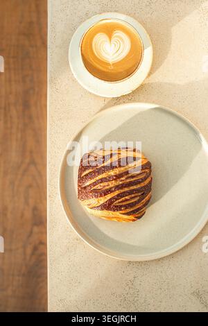 Ein Blick von oben auf ein frisches zweifarbiges Schokoladen-Croissant-Gebäck auf einer Keramikplatte, serviert zusammen mit einer heißen Tasse Latte Art Kaffee. Stockfoto