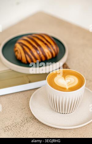 Ein minimalistischer Blick von oben auf ein frisches, individuelles Babka-Gebäck mit Schokoladenswirl auf einer dunkelgrünen Keramikplatte, serviert neben einer heißen Tasse Latte Art Cof Stockfoto