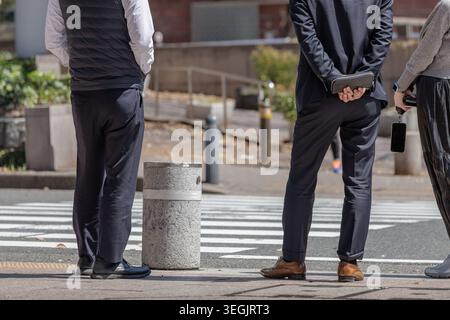 Mittags – drei Kollegen warten an einem Crosswalk, Hände hinter dem Rücken halten eine Clutch-Geldbörse oder Telefone in der Hand und führen zwanglose Gespräche. Stockfoto