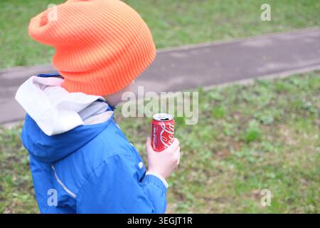 Ein Kind in einer hellblauen Jacke und einem orangefarbenen Hut hält eine Coca-Cola-Dose, während es draußen in der Nähe eines Wohngebäudes spaziert. Moskau, Russland, 04.23-2026 Stockfoto