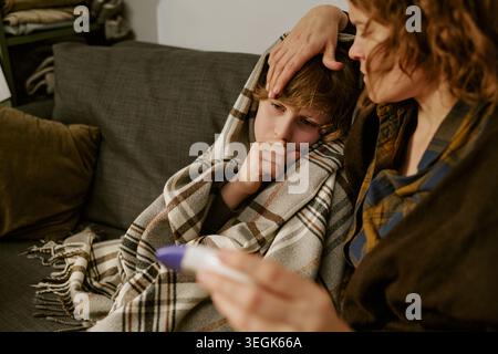 Mutter tröstet krankes Kind, während sie das Thermometer zu Hause überprüft Stockfoto