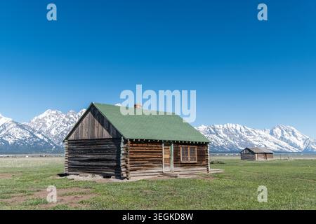 Historische Blockhütte in einem riesigen grünen Feld mit majestätischen schneebedeckten Bergen unter einem klaren blauen Himmel. Ruhig und malerisch. Ideal für Reisen, Natur, Stockfoto