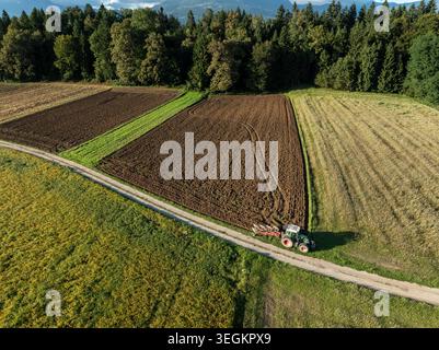Der Traktor arbeitet auf gepflügten Feldern in der Nähe eines Waldes. Berge sind im Hintergrund. Bei dieser Aktivität wird die Landwirtschaft tagsüber gezeigt. Stockfoto