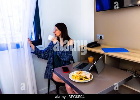 Frau, die die Vorhänge öffnet und im Hotelzimmer frühstückt Stockfoto