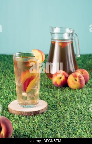 Ein Glas eiskalten Pfirsichtee auf einer hölzernen Achterbahn mit einem Krug und ganzen Früchten im Hintergrund auf grünem Gras Stockfoto