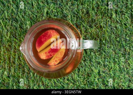 Blick auf eine Glaskanne mit pfirsichfarbenem Eistee und Obstscheiben auf einem leuchtend grünen Rasen Stockfoto