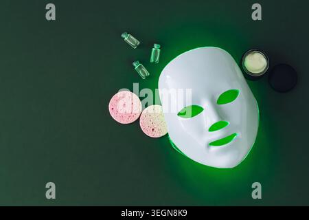 Trendige Hautpflege-Komposition mit futuristischer LED-Gesichtsmaske und Beauty-Accessoires auf dunkelgrünem Hintergrund. Luxuriöse Hautpflege und Beauty-Konzept. Stockfoto
