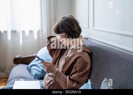 Die nachdenkliche verzögerte Millennial-Frau sitzt auf dem Sofa mit Smartphone, berührt den Hals mit leichten Unannehmlichkeiten, indem sie soziale Medien durchblättert und sich online müde fühlt Stockfoto