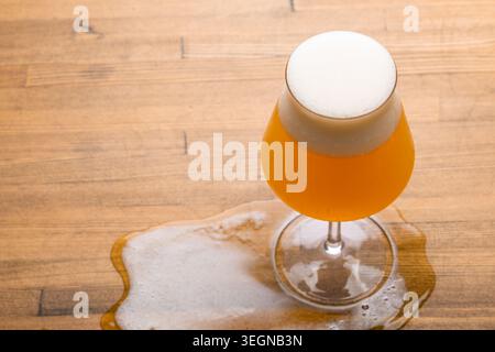 Tulpenglas mit trübem Bernsteinbier mit dichtem Schaum und einem Auslauf über eine hölzerne Tischplatte, von oben gesehen Stockfoto