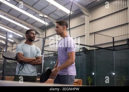 Verschiedene männliche Freunde stehen und unterhalten sich neben dem Paddelplatz in Sportbekleidung und halten Paddel Stockfoto