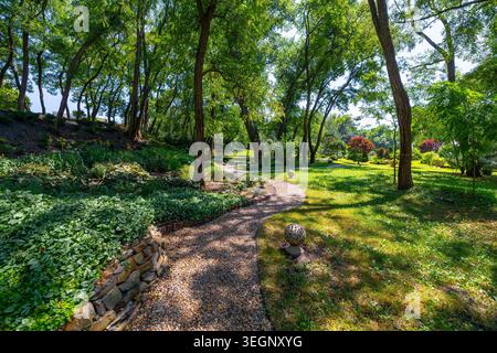 Ein gewundener Kiesweg führt durch einen öffentlichen Park, der von hohen grünen Laubbäumen, grünen Rasenflächen und Gartenbeeten mit ston umgeben ist Stockfoto