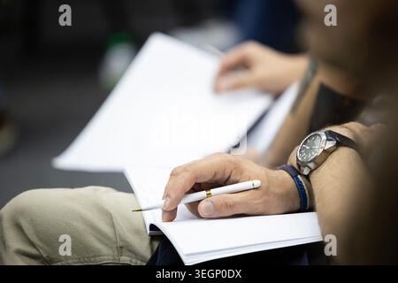 Nahaufnahme eines Mannes, der einen weißen Kugelschreiber über ein offenes Notizbuch hält, während er mit anderen Teilnehmern an einem Geschäftsseminar oder einer Konferenz teilnimmt Stockfoto