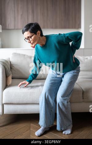 Ältere Frau mit Wirbelsäulenerkrankung, die nach dem Aufstehen vom Sofa an plötzlichen Rückenschmerzen leidet. Stockfoto