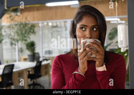 Indische Frau, die im Büro einen gerippten Becher in der Nähe des Mundes hält, trägt ein burgunderrotes Oberteil, Smartwatch, Kopierraum Stockfoto