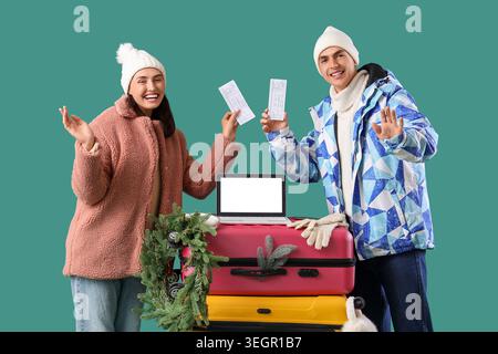 Nettes junges Paar mit Koffern, Tickets, Laptop und Weihnachtsaccessoires auf grünem Hintergrund. Winterurlaubskonzept Stockfoto