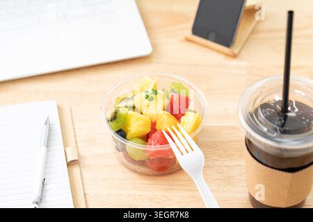 Eine Schüssel mit Obstsalat liegt auf einem Schreibtisch zwischen Laptop, Notebook und Eiskaffee und ist bereit für einen anstrengenden Arbeitstag. Stockfoto