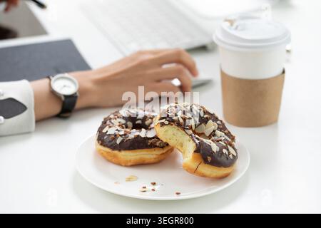 Eine Person sitzt an einem weißen Schreibtisch, arbeitet an einem Laptop, während sie einen in Schokolade getauchten Donut und eine Tasse Kaffee genießt. Stockfoto