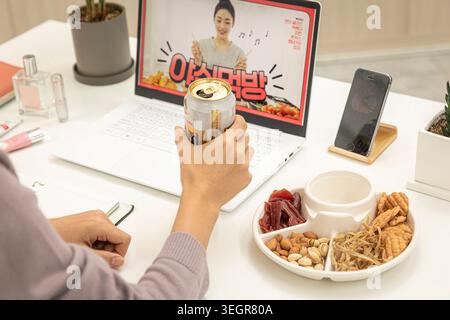 Eine Frau sitzt an einem Schreibtisch, hält eine Dose Bier und sieht sich eine Kochshow auf ihrem Laptop an. Stockfoto